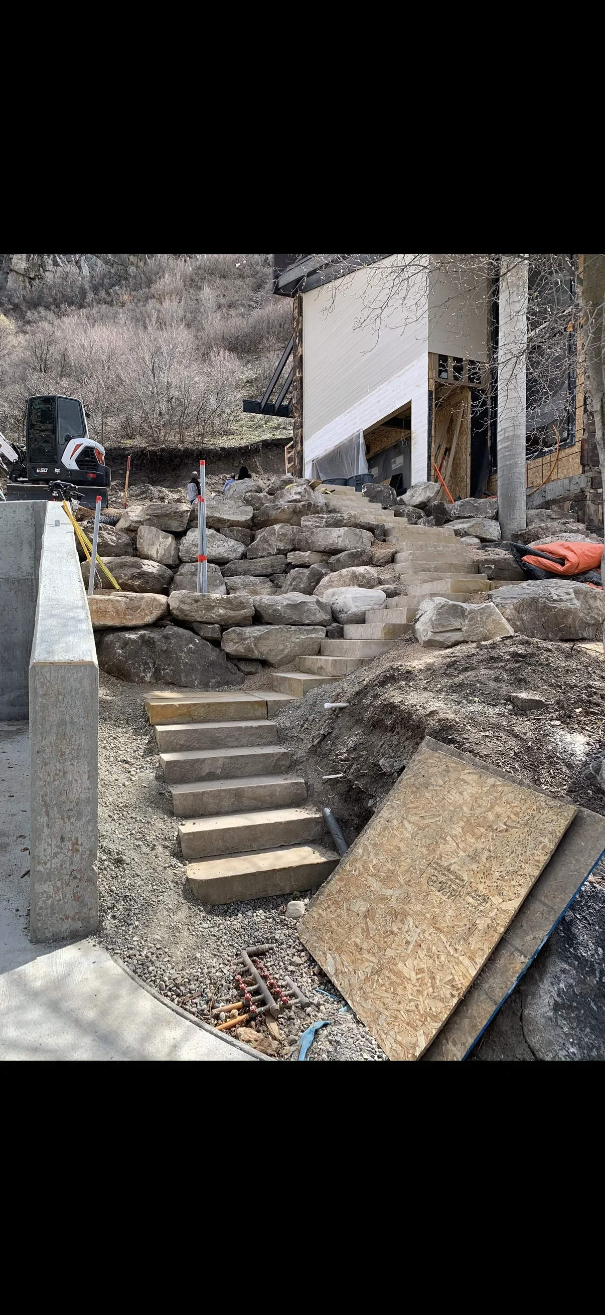 Large boulder retaining wall with cut stone steps climbing a graded slope beside a building under construction