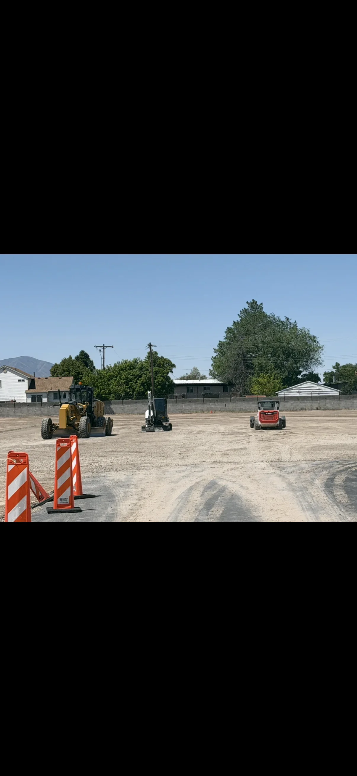 Motor grader, mini excavator, and skid steer grading a gravel site with orange barricades in the foreground