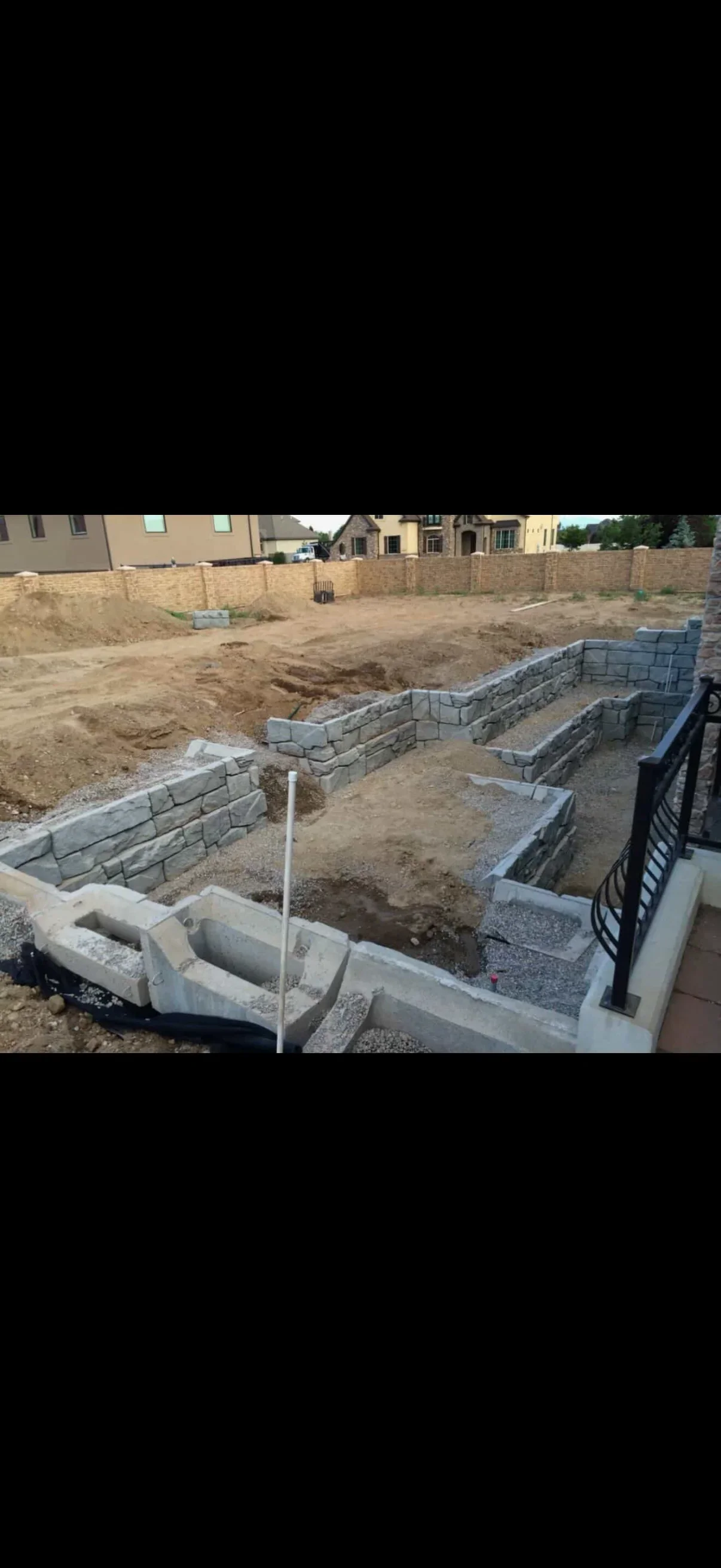 Tiered gray split-face concrete retaining walls on gravel base with wrought iron railing and excavated site