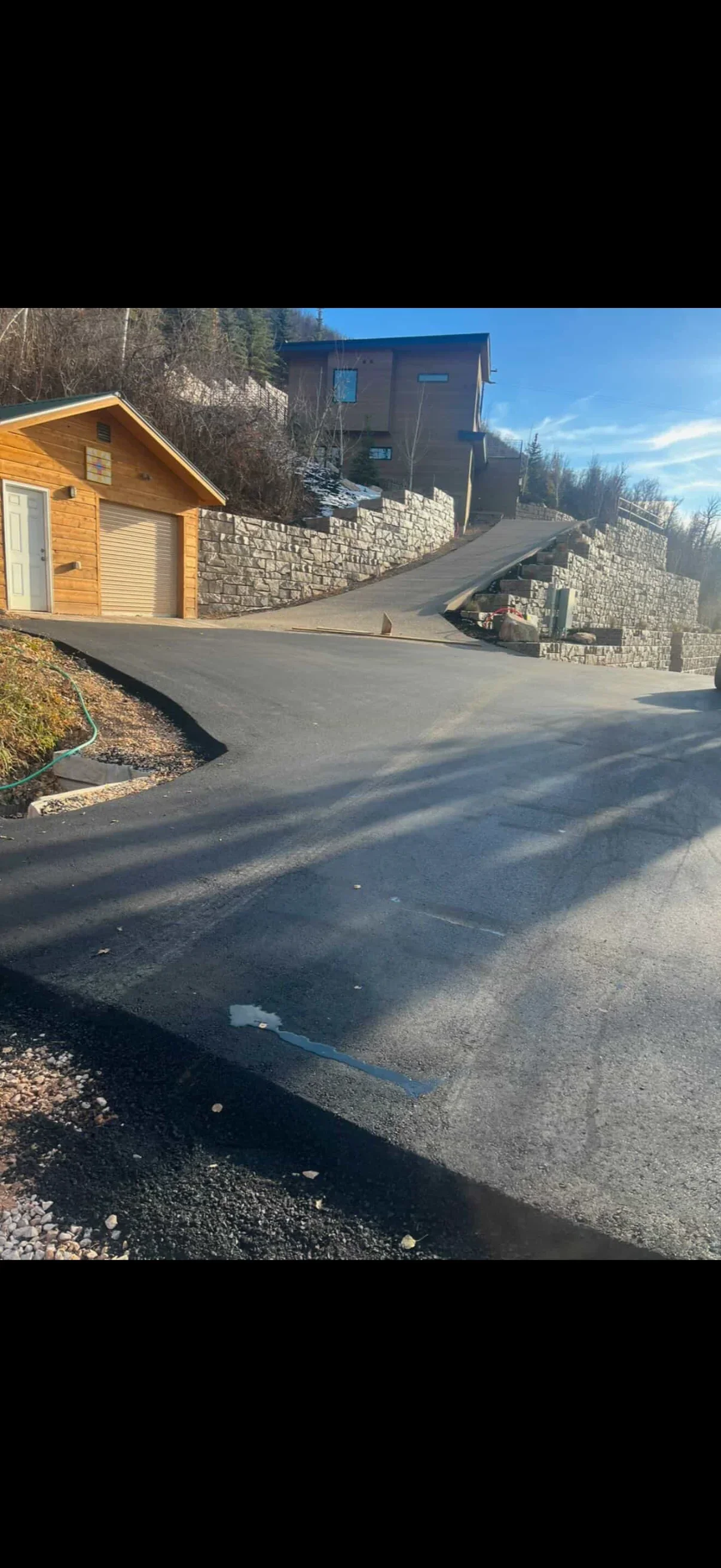 Newly paved asphalt driveway with gray segmented stone retaining walls on a hillside next to a log-style garage