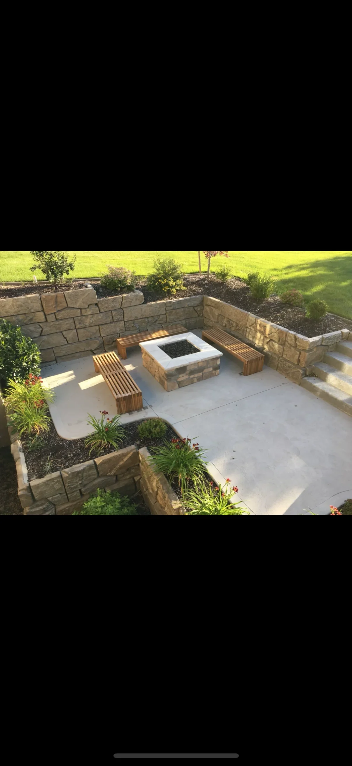 Tan stacked stone retaining walls, concrete patio, stone fire pit, and wooden slat benches with mulched planting beds