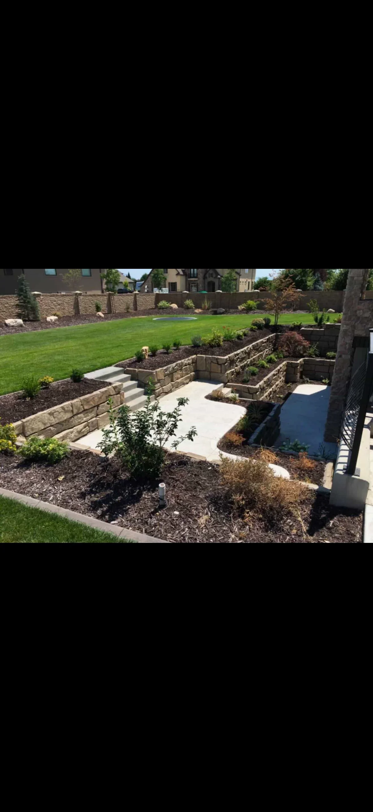 Tiered tan segmented stone retaining walls with concrete steps, curved patio, and mulched planting beds on a sloped backyard