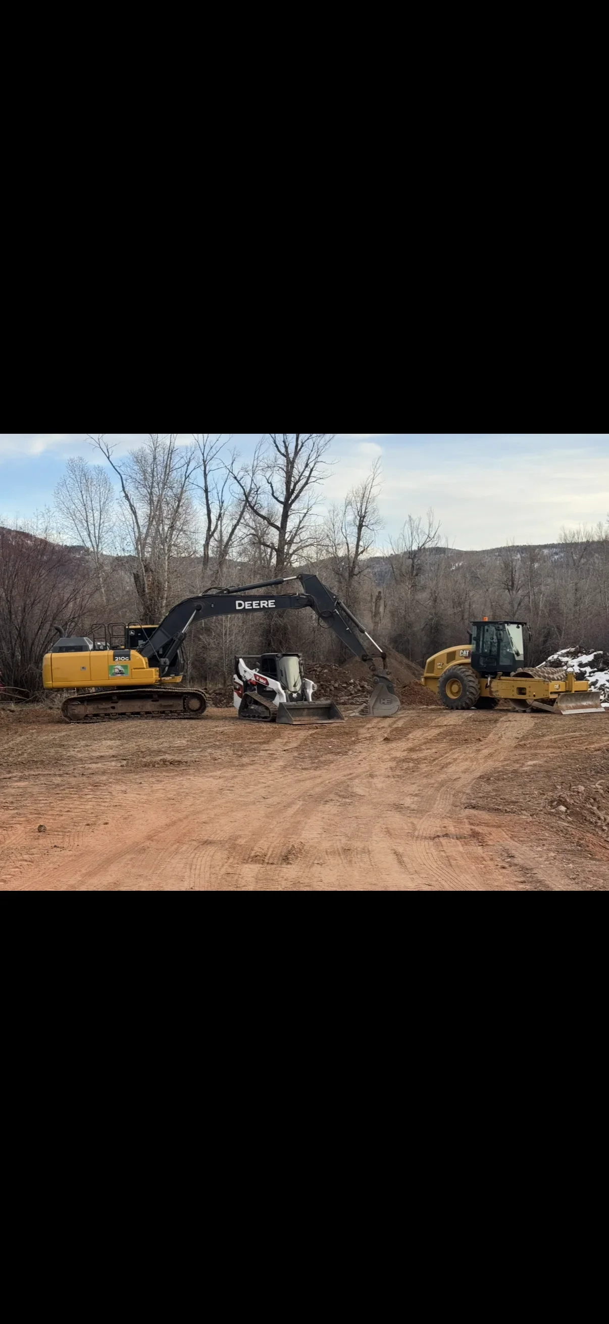 John Deere 210G excavator, Bobcat compact track loader, and Caterpillar compactor grading bare dirt site
