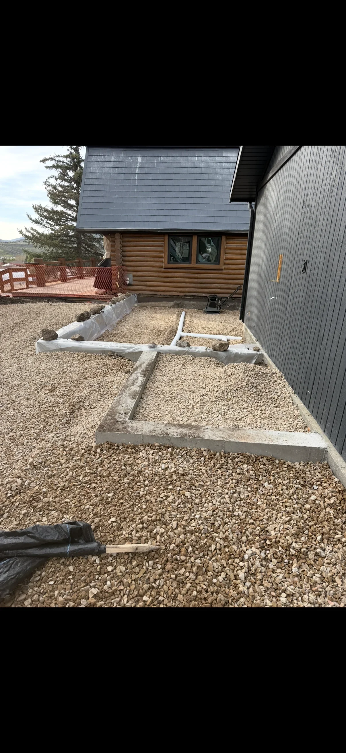 Concrete footing forms set in crushed gravel with white PVC pipe and a plate compactor beside a log home foundation