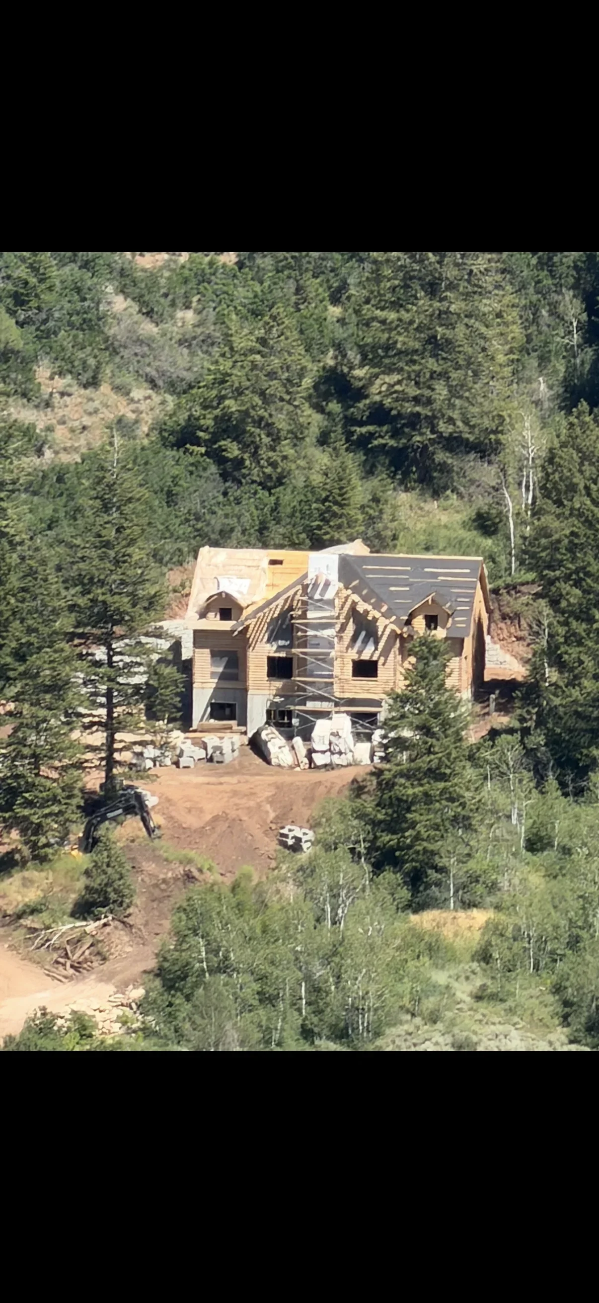 Partially framed timber home with dormers and roof underlayment on a graded hillside, stacked stone and excavator on site
