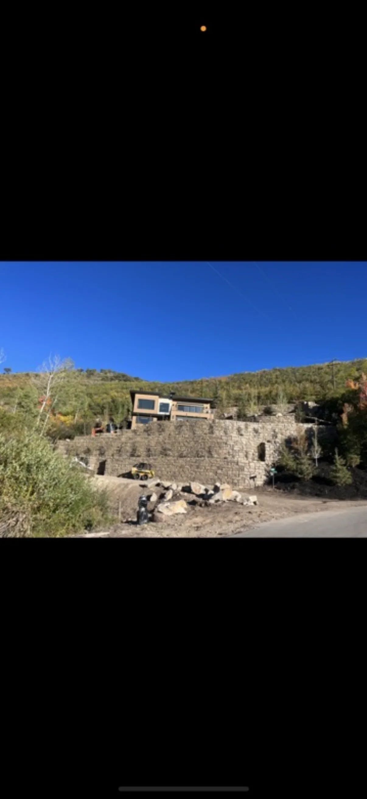 Large irregular natural stone retaining wall on a graded hillside with excavated boulders and a compact track loader